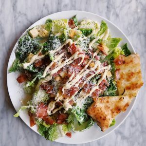 Tasty Caesar salad topped with bacon, cheese, and drizzled sauce, served with crispy bread.